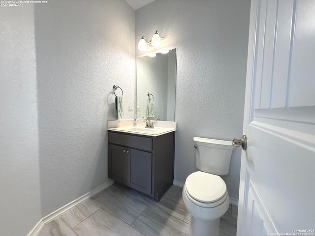 half bath featuring toilet, a textured wall, baseboards, and vanity