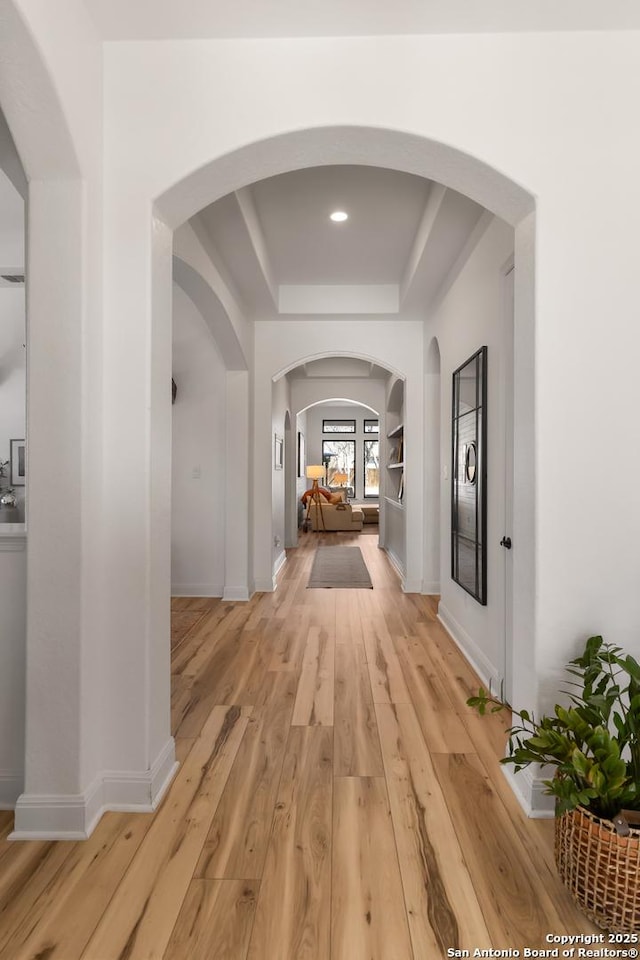 corridor with arched walkways, a tray ceiling, light wood-style flooring, and baseboards