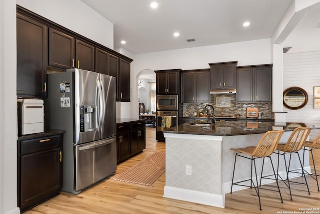 kitchen with light wood finished floors, arched walkways, appliances with stainless steel finishes, dark stone countertops, and a sink