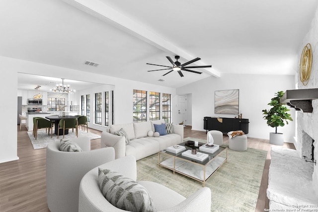 living room with visible vents, a fireplace with raised hearth, wood finished floors, vaulted ceiling with beams, and ceiling fan with notable chandelier