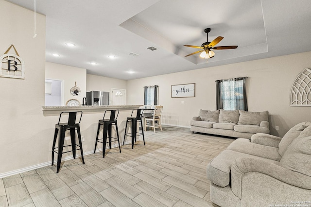 living area with ceiling fan, wood finish floors, visible vents, baseboards, and a tray ceiling