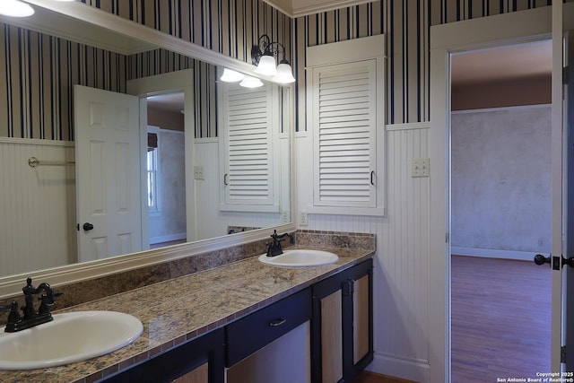 bathroom with double vanity, wainscoting, a sink, and wallpapered walls