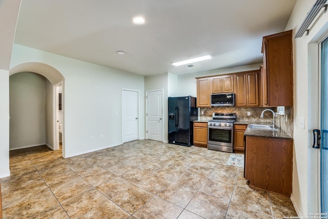 kitchen with arched walkways, stainless steel appliances, a sink, backsplash, and brown cabinetry