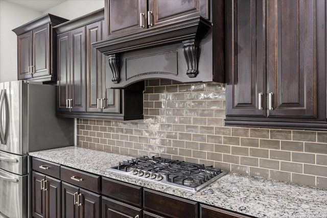 kitchen featuring light stone counters, stainless steel appliances, dark brown cabinets, custom exhaust hood, and tasteful backsplash