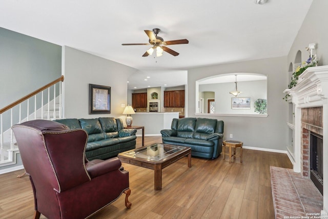 living area with baseboards, hardwood / wood-style flooring, ceiling fan, stairs, and a brick fireplace