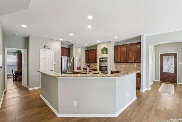 kitchen with arched walkways, wood finished floors, appliances with stainless steel finishes, a large island, and tasteful backsplash