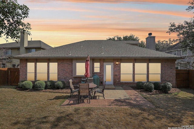 back of property at dusk featuring a patio, brick siding, fence, a lawn, and roof with shingles