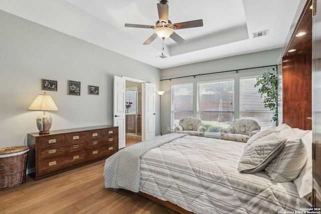 bedroom with a tray ceiling, visible vents, ensuite bathroom, a ceiling fan, and light wood-type flooring