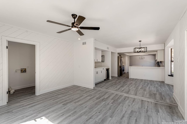 unfurnished living room with ceiling fan with notable chandelier, light wood-type flooring, visible vents, and baseboards