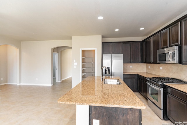 kitchen with arched walkways, stainless steel appliances, a kitchen island with sink, a sink, and light stone countertops