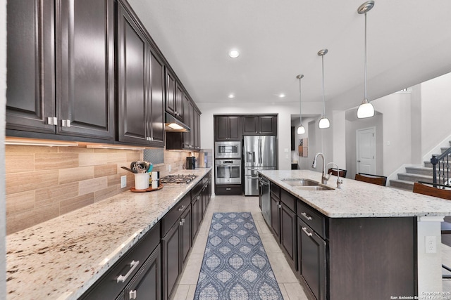 kitchen with a center island with sink, backsplash, appliances with stainless steel finishes, a sink, and light stone countertops