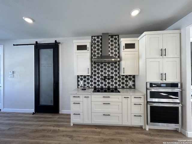 kitchen featuring a barn door, stainless steel double oven, black electric cooktop, wall chimney range hood, and tasteful backsplash