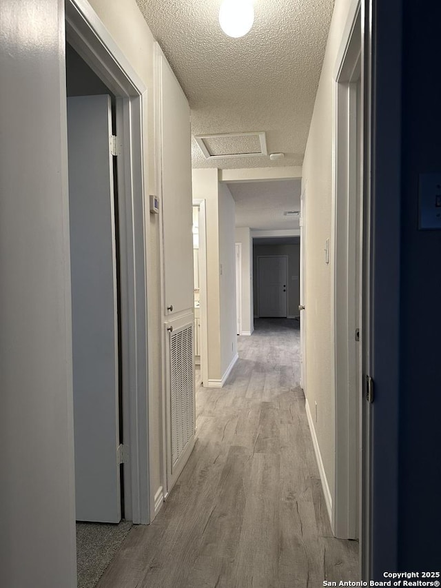 hall with light wood finished floors, attic access, baseboards, and a textured ceiling