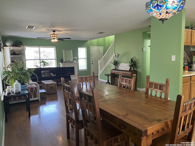 dining area with ceiling fan, dark wood-style flooring, a fireplace, visible vents, and stairs