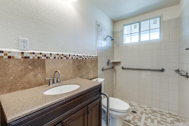 bathroom featuring tile walls, a textured wall, toilet, vanity, and tiled shower