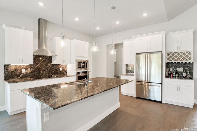 kitchen with stainless steel appliances, wood finished floors, a sink, wall chimney exhaust hood, and a center island with sink