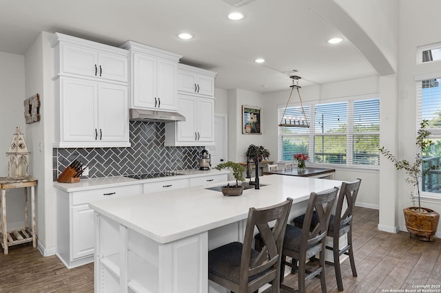 kitchen with a kitchen island with sink, black electric cooktop, under cabinet range hood, wood finished floors, and light countertops