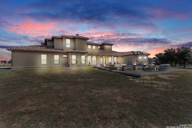 back of house at dusk featuring a tiled roof, a lawn, a patio, and stucco siding