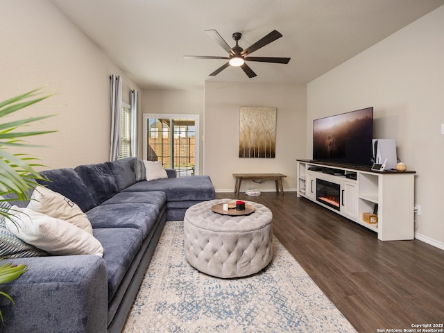 living room featuring a ceiling fan, a glass covered fireplace, dark wood-style flooring, and baseboards