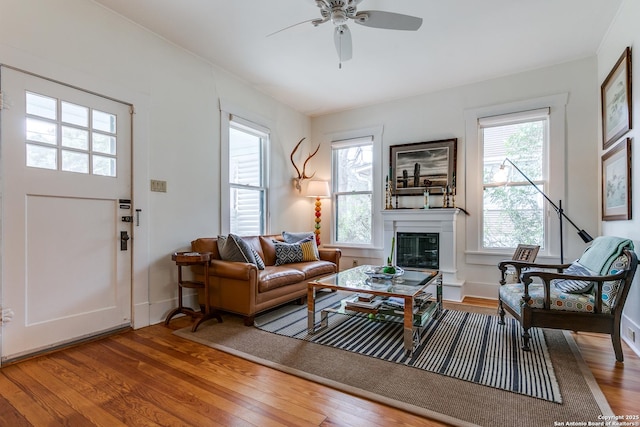 living area featuring a ceiling fan, a glass covered fireplace, baseboards, and wood finished floors