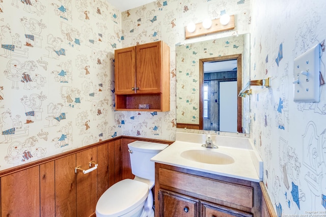 bathroom with toilet, wallpapered walls, vanity, and wainscoting