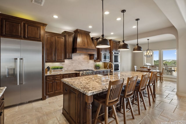 kitchen featuring built in appliances, stone tile floors, premium range hood, visible vents, and tasteful backsplash