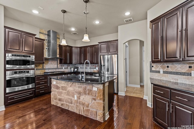 kitchen with arched walkways, stainless steel appliances, visible vents, a sink, and wall chimney range hood