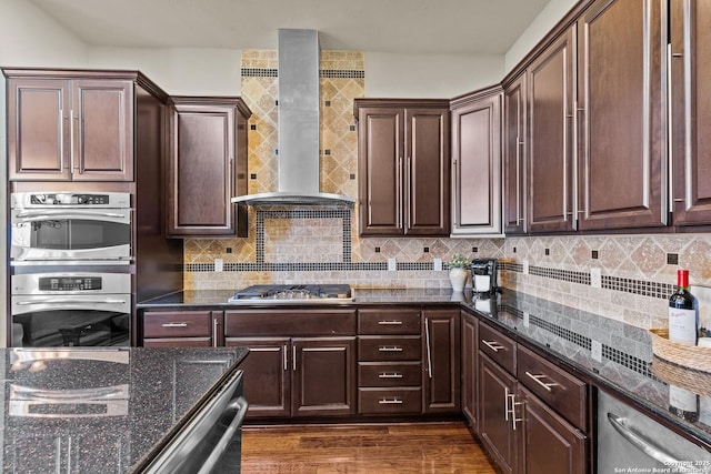 kitchen with appliances with stainless steel finishes, wall chimney exhaust hood, tasteful backsplash, dark stone countertops, and dark wood finished floors