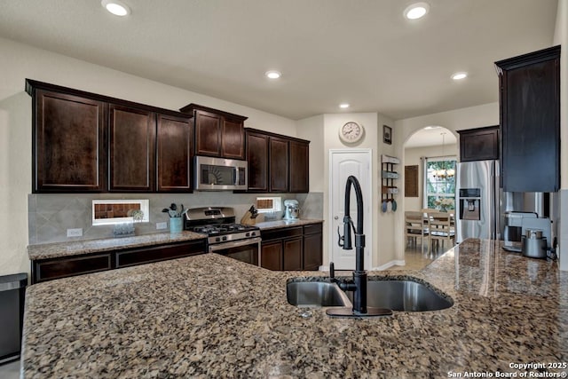 kitchen with arched walkways, appliances with stainless steel finishes, a sink, dark stone countertops, and dark brown cabinets