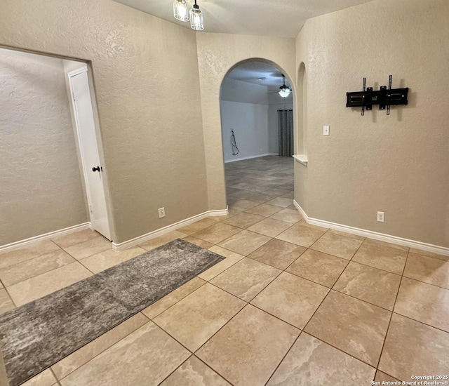 tiled empty room featuring baseboards, arched walkways, ceiling fan, and a textured wall