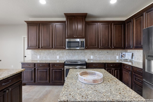 kitchen featuring backsplash, appliances with stainless steel finishes, dark brown cabinetry, a textured ceiling, and light stone countertops