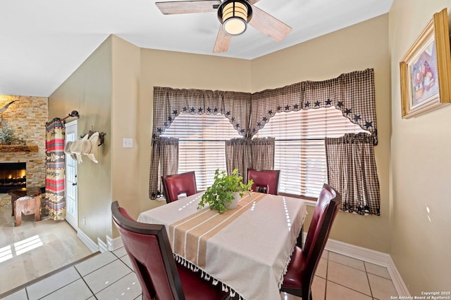 tiled dining area with ceiling fan, a stone fireplace, and baseboards