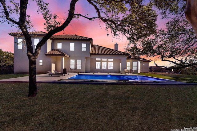 back of house at dusk with a patio, a tiled roof, a lawn, an outdoor pool, and a chimney