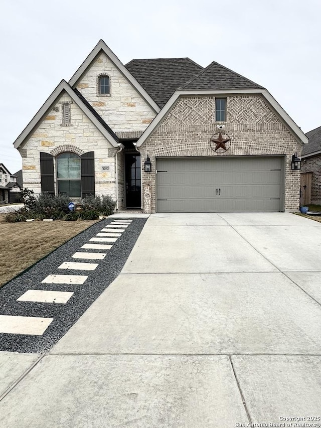 french country inspired facade with brick siding, driveway, an attached garage, and roof with shingles