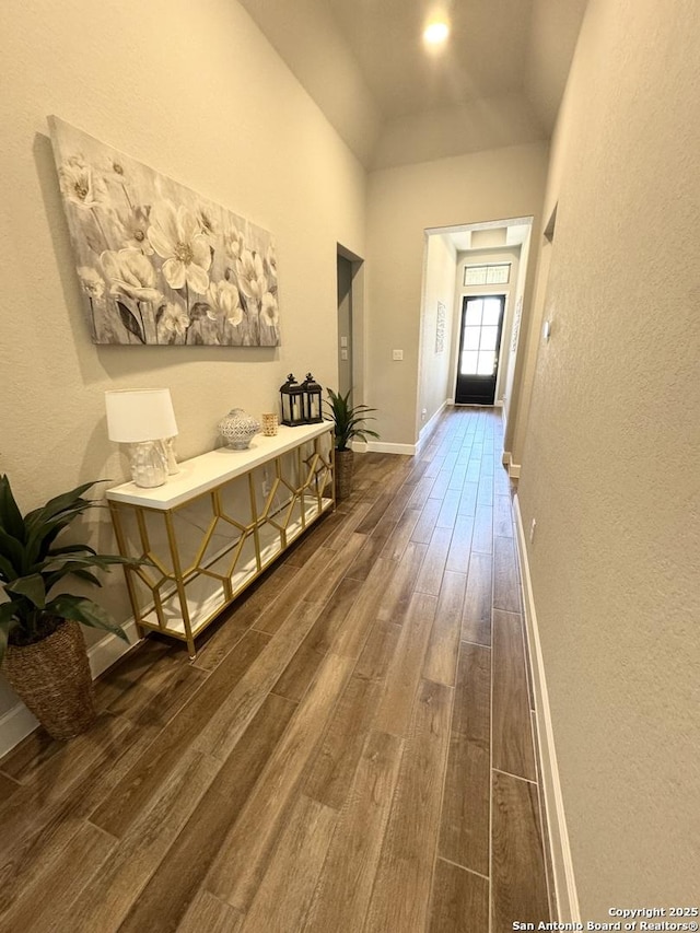 corridor with dark wood-type flooring, a textured wall, vaulted ceiling, and baseboards