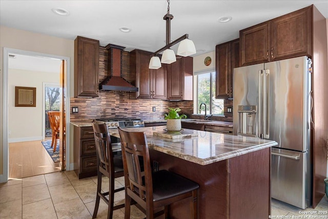 kitchen featuring wall chimney exhaust hood, light stone counters, backsplash, high end fridge, and a sink