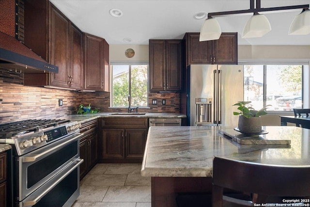 kitchen featuring decorative backsplash, appliances with stainless steel finishes, dark brown cabinetry, a sink, and wall chimney exhaust hood