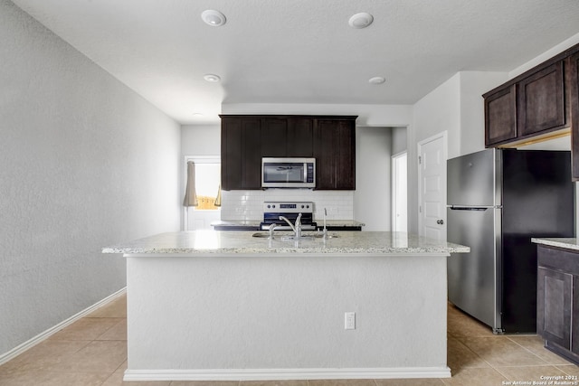 kitchen with light stone counters, light tile patterned floors, stainless steel appliances, backsplash, and a kitchen island with sink