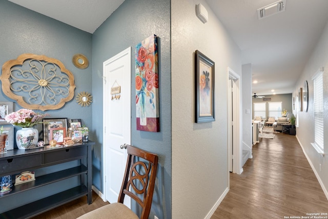 hall with a textured wall, wood finished floors, visible vents, and baseboards