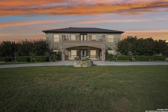 view of front facade with driveway, a balcony, stone siding, metal roof, and a yard