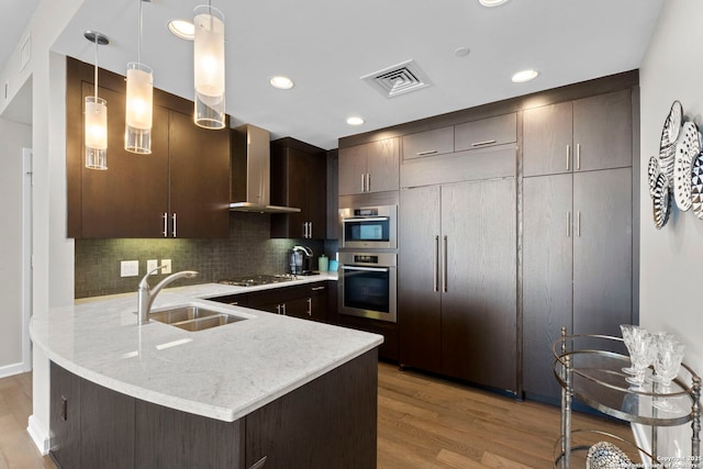 kitchen featuring a peninsula, a sink, visible vents, dark brown cabinets, and wall chimney exhaust hood