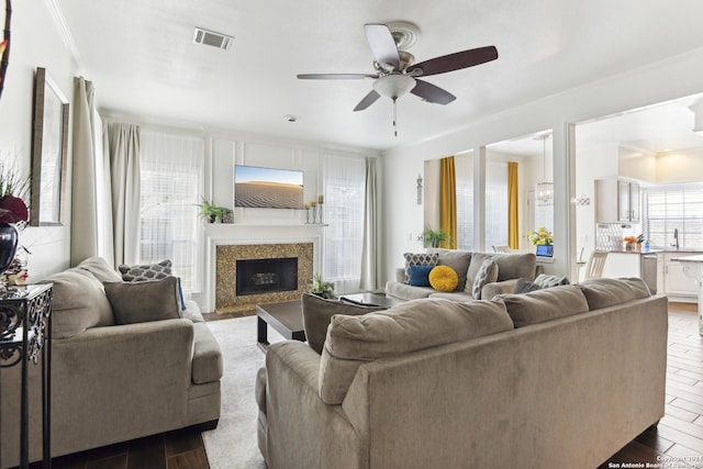 living area with dark wood-type flooring, a fireplace, visible vents, a ceiling fan, and ornamental molding