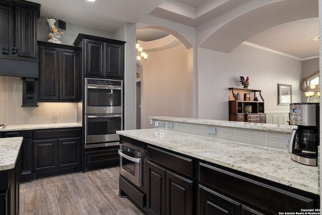 kitchen featuring double oven, light stone counters, dark cabinetry, light wood finished floors, and crown molding