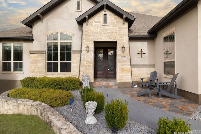 view of exterior entry featuring stone siding, french doors, a shingled roof, and stucco siding