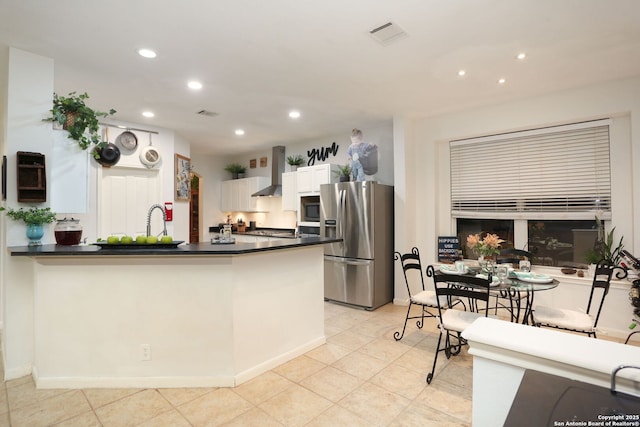 kitchen featuring stainless steel appliances, dark countertops, white cabinets, a peninsula, and wall chimney exhaust hood