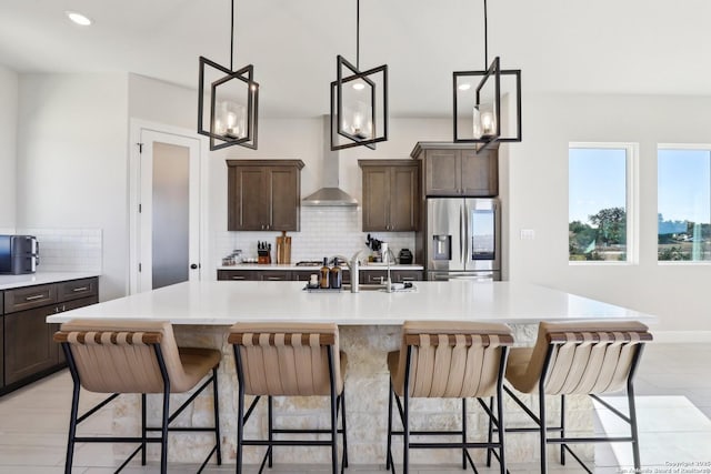 kitchen featuring a center island with sink, light countertops, decorative backsplash, dark brown cabinetry, and stainless steel fridge