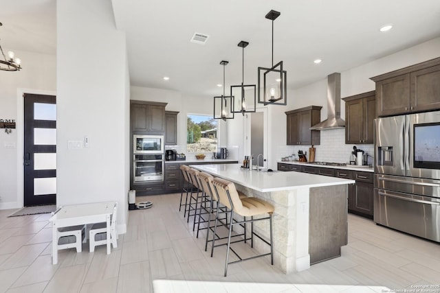 kitchen featuring dark brown cabinetry, a breakfast bar, appliances with stainless steel finishes, decorative backsplash, and wall chimney exhaust hood