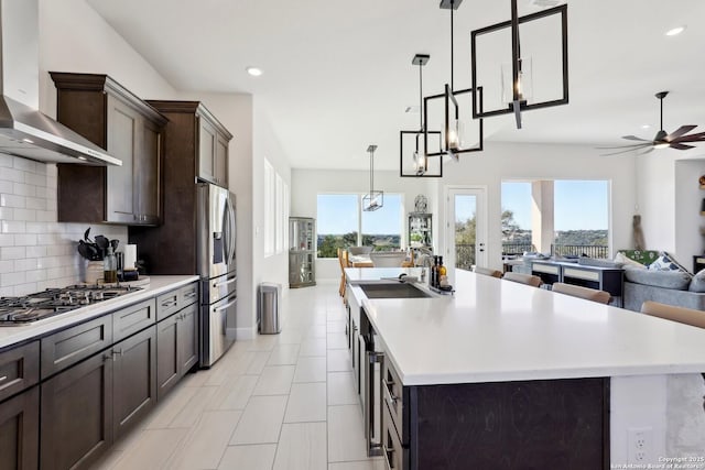 kitchen with dark brown cabinetry, wall chimney exhaust hood, appliances with stainless steel finishes, open floor plan, and light countertops