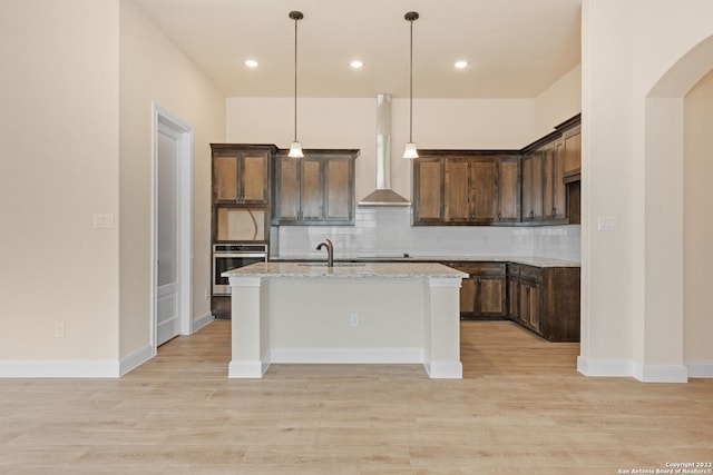 kitchen featuring backsplash, a kitchen island with sink, a sink, wall chimney range hood, and oven