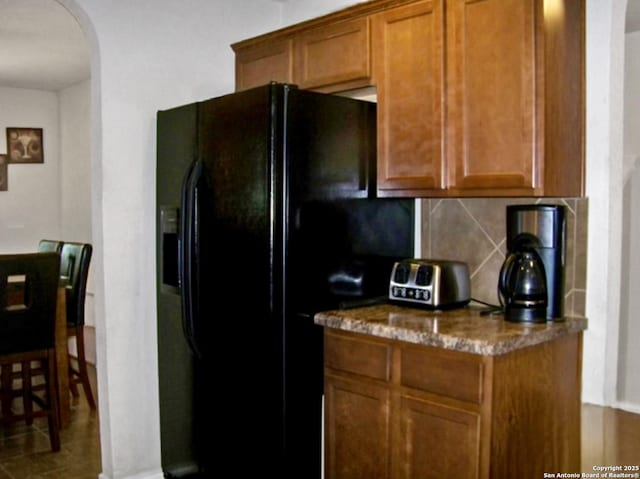 kitchen with arched walkways, black fridge with ice dispenser, decorative backsplash, brown cabinetry, and dark stone countertops